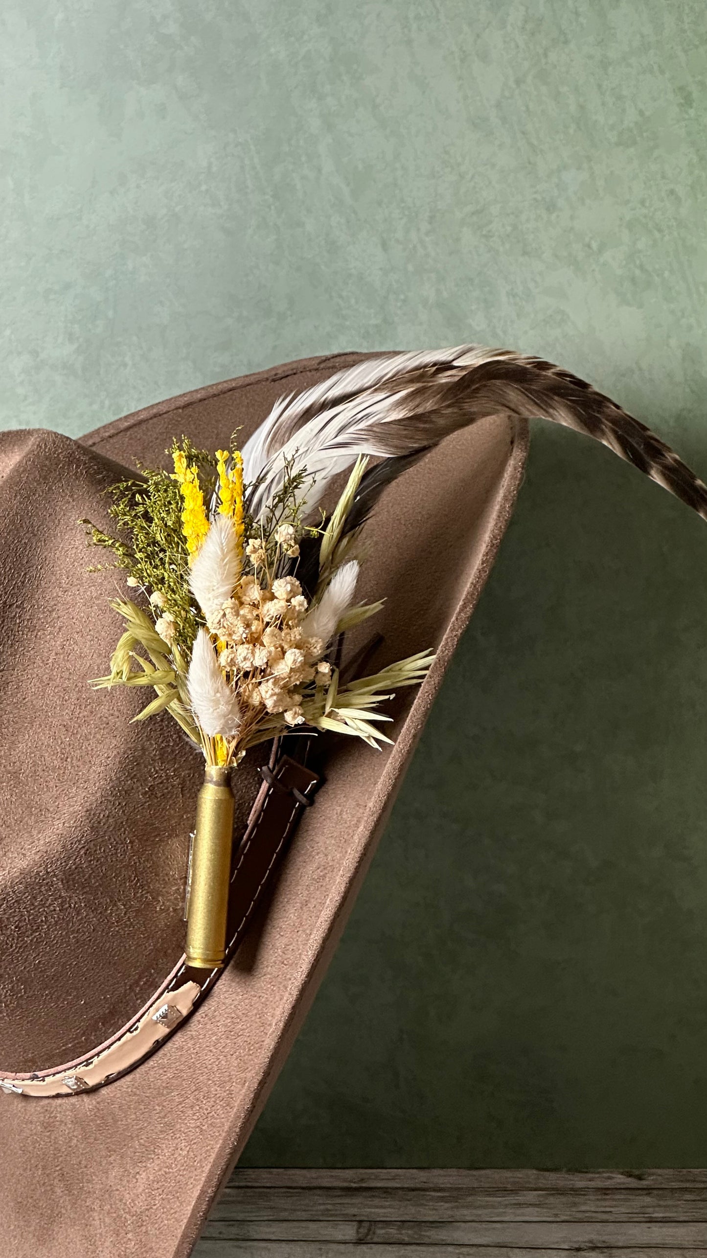 A brown cowboy hat with a decorative dried flower boutonniere featuring a gold bullet casing, white feathers, and yellow accents, set against a textured sage green background.