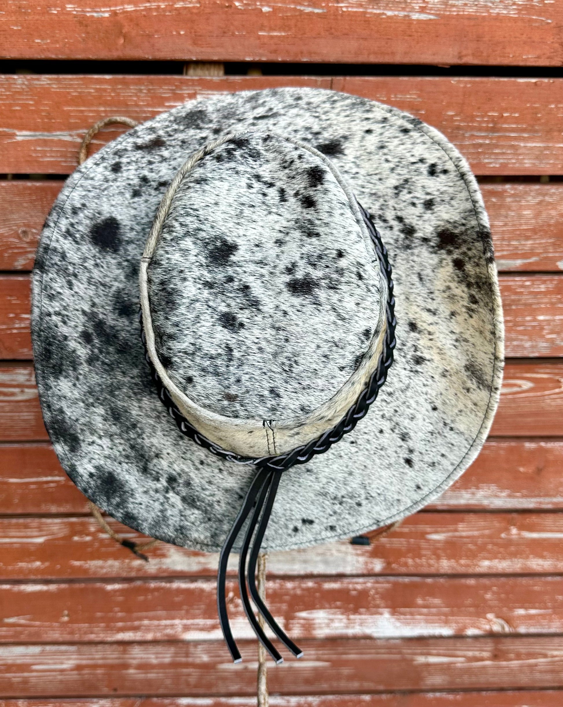 Goat hide hat with black speckles and braided leather band on a wooden surface