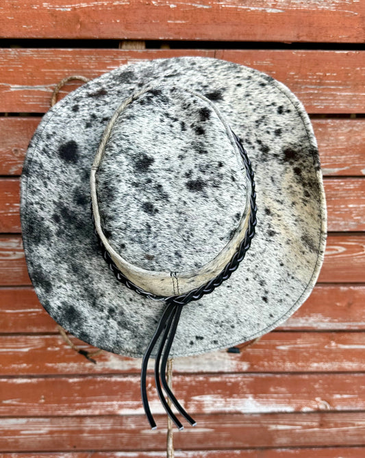Goat hide hat with black speckles and braided leather band on a wooden surface