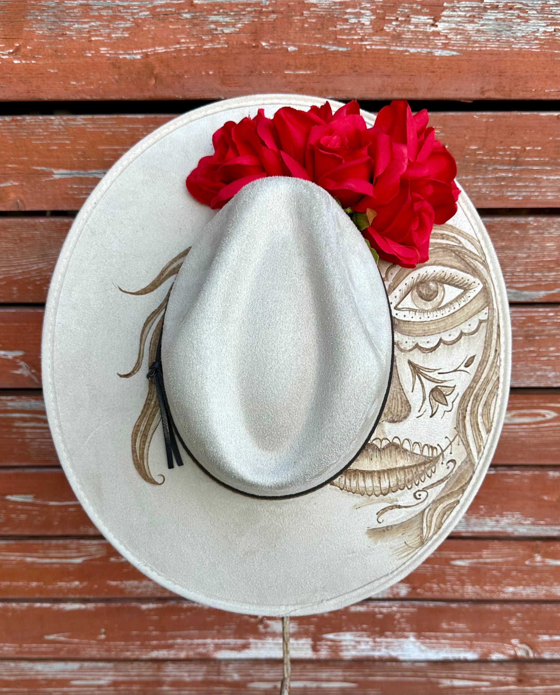 White cowboy hat with Handburned Catrina decorated with red roses on a wooden surface