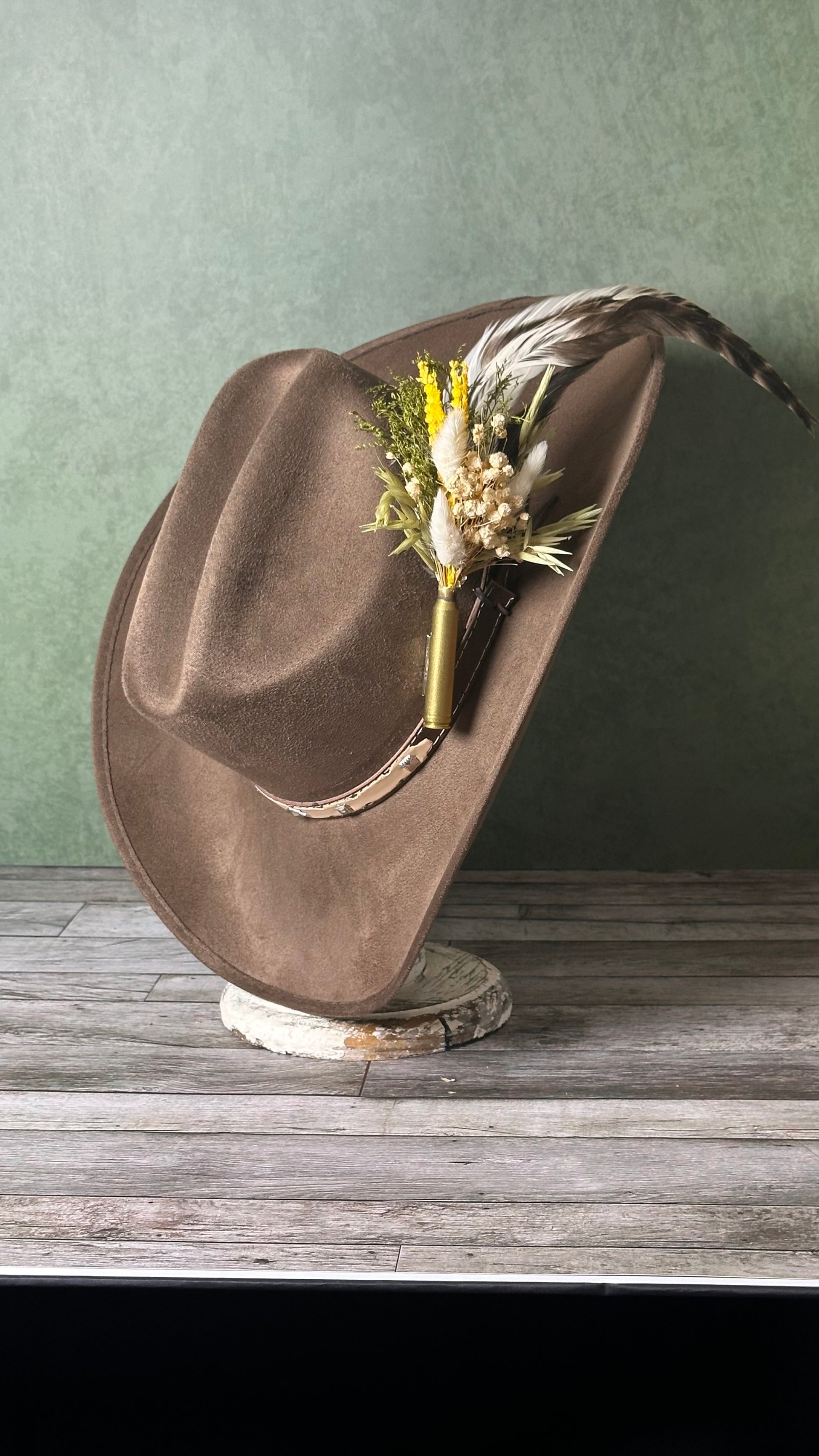 A brown cowboy hat displayed on a rustic wooden stand against a textured green wall. The hat features a decorative dried floral and feather accent on the crown.